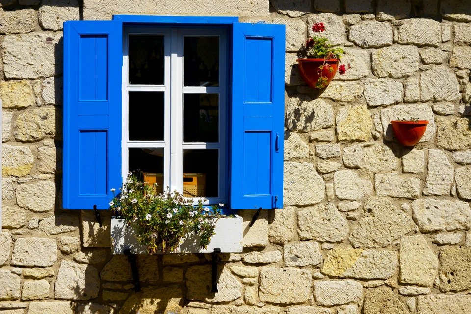 Fa&ccedil;ade en pierre traditionnelle avec fen&ecirc;tre &agrave; volets bleus ouverts, encadrement blanc, jardini&egrave;re avec plantes fleuries sur rebord