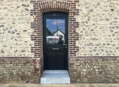Façade extérieure d'habitation avec porte d'entrée noire à panneaux et vitrage, encadrée par un arc en briques rouges