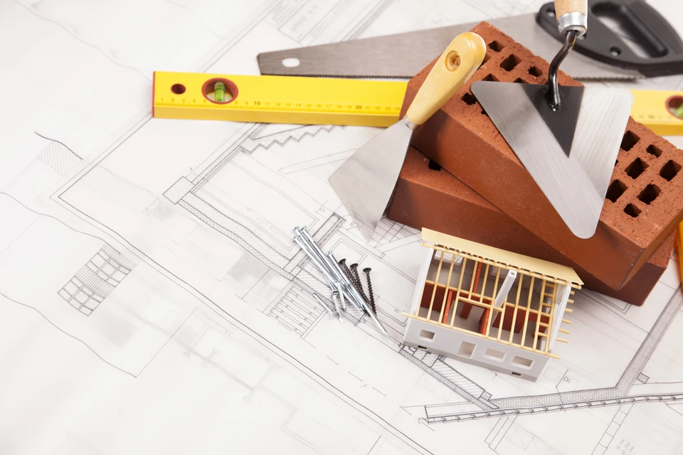 Chantier de construction extérieur avec maçon en salopette bleue et t-shirt rouge tenant une brique perforée et une truelle