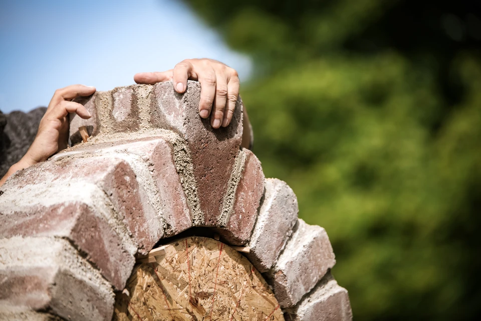 Homme d'&acirc;ge m&ucirc;r aux cheveux grisonnants portant un sweat-shirt gris fonc&eacute;, photographi&eacute; de dos en train de mesurer ou d'&eacute;valuer un mur de briques